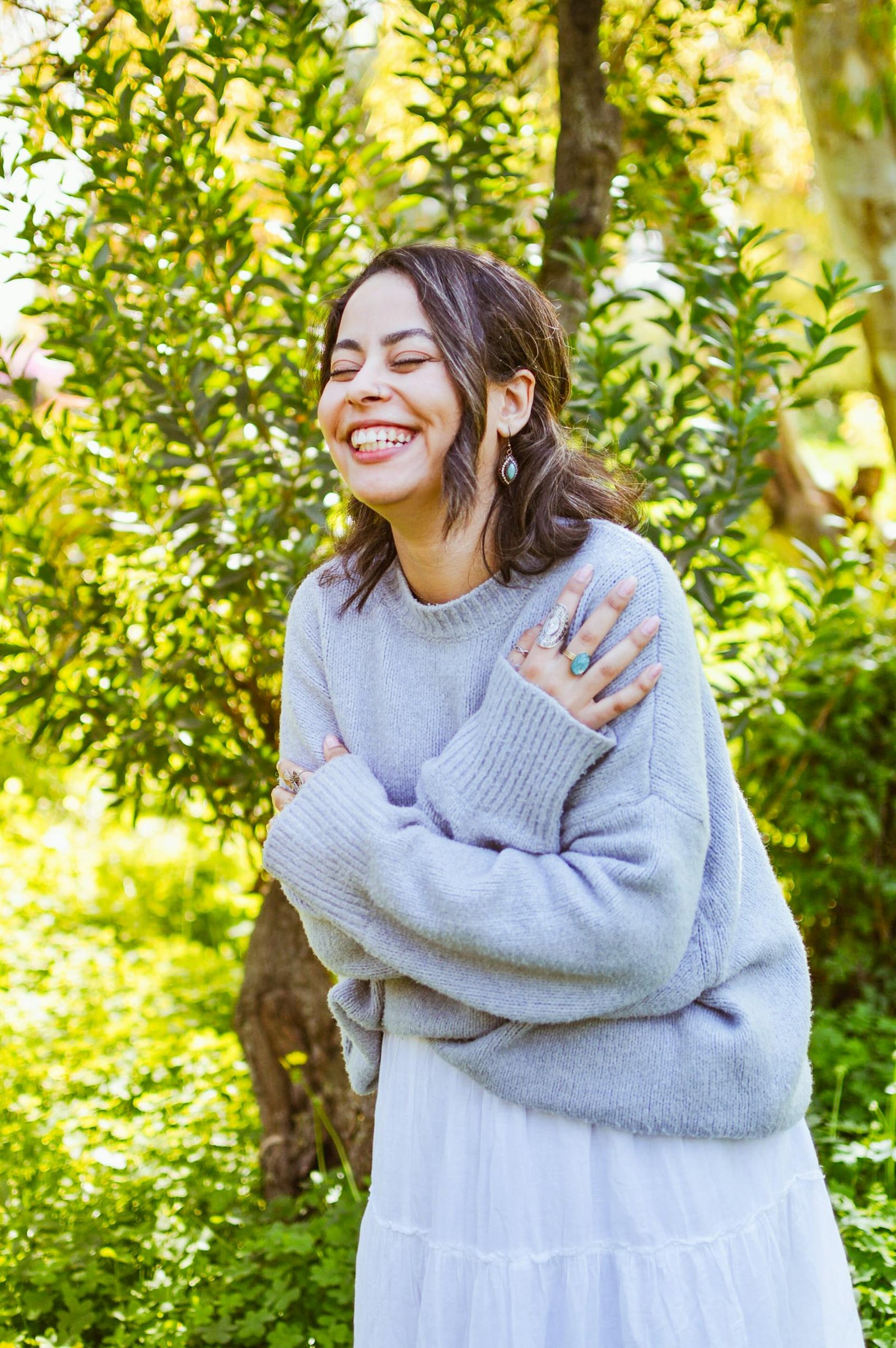 Smiling woman in a sweater embraces herself in a sunny garden.